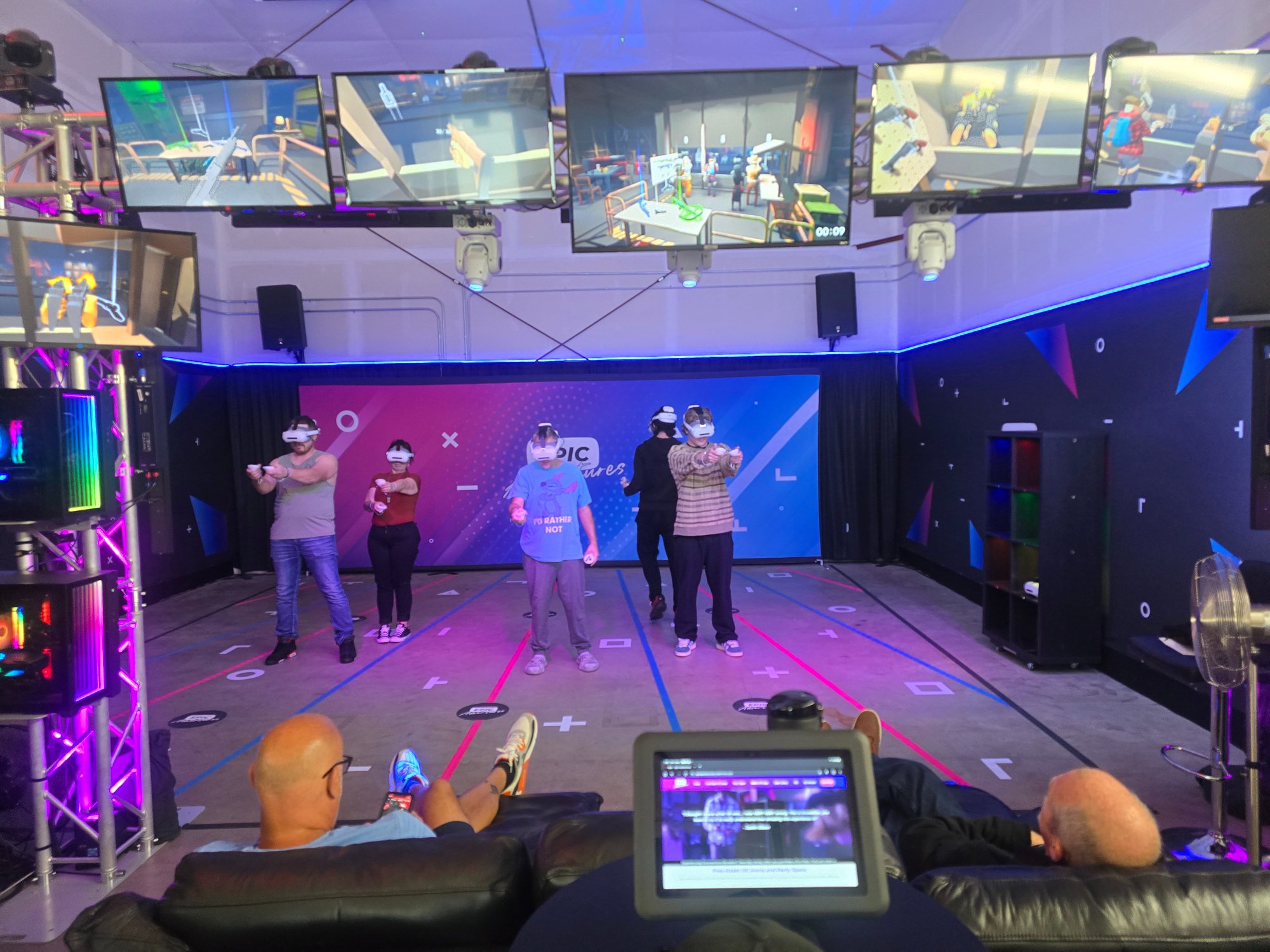 Five players in VR headsets with spectators watching from the couch and game screens overhead at Epic Adventures