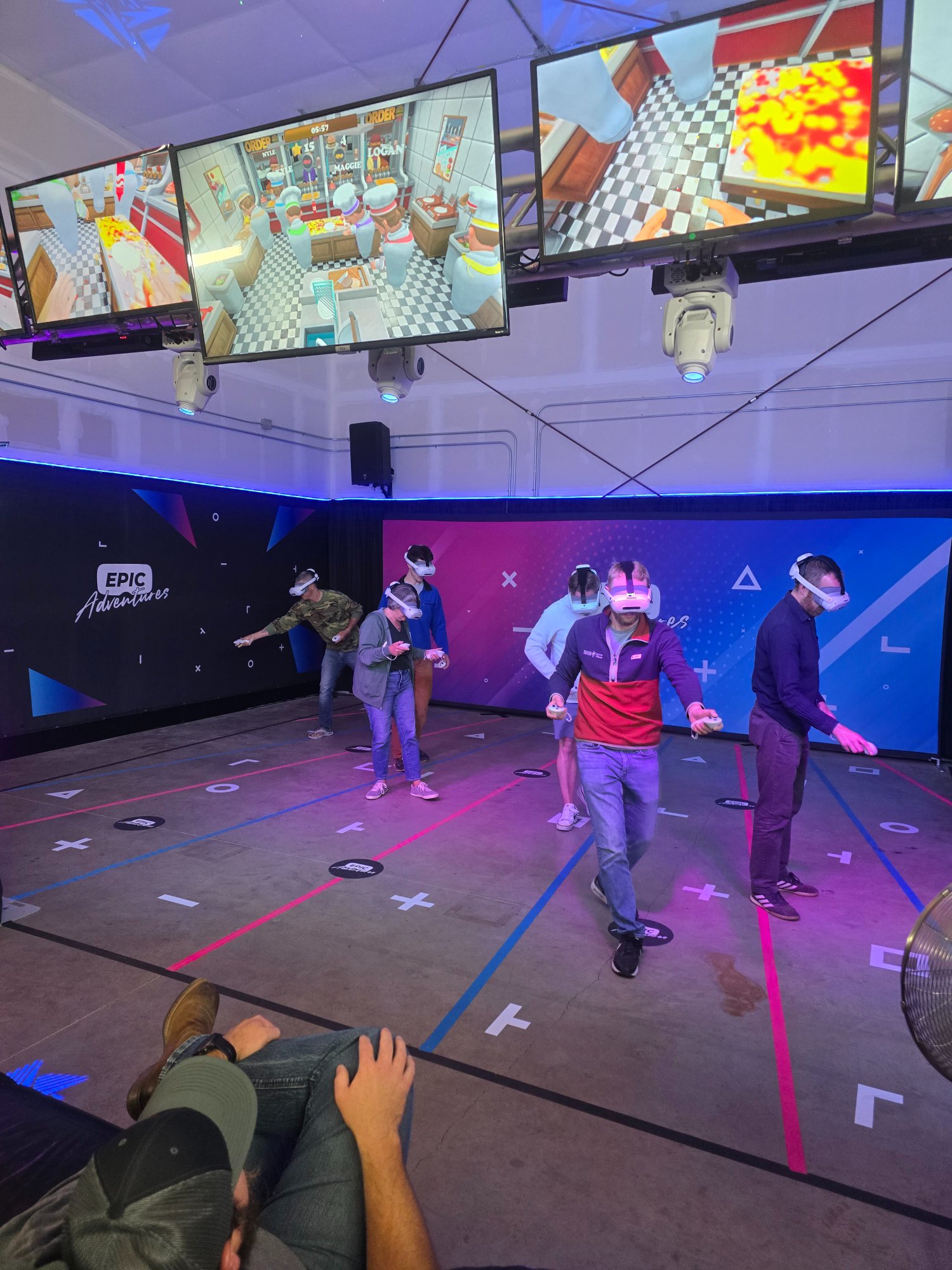 Corporate team playing Cook'd Up VR cooking game together with spectator screens showing the in-game action at Epic Adventures